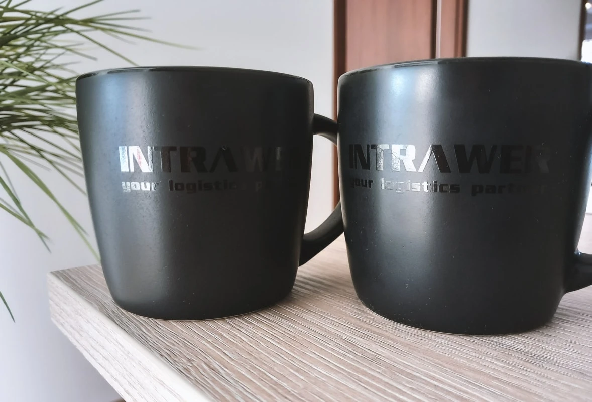 stojące na beżowym blacie, dwa czarne kubki z nadrukowanym napisem INTRAWER, z widocznymi liśćmi w tle